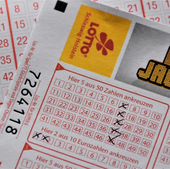 A close-up image of lottery tickets featuring crossed-out numbers, brand logos, and various numbers in a grid format. The tickets have sections for different games, and numbers are marked with black crosses.