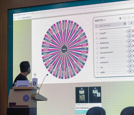 A person stands in front of a large screen displaying a colorful digital wheel with names. The screen shows a web interface with options for inputs, featuring a list of names with checkboxes on one side and a large, vibrant spinning wheel on the other. The environment appears to be indoors, possibly in a conference or presentation setting with a podium.